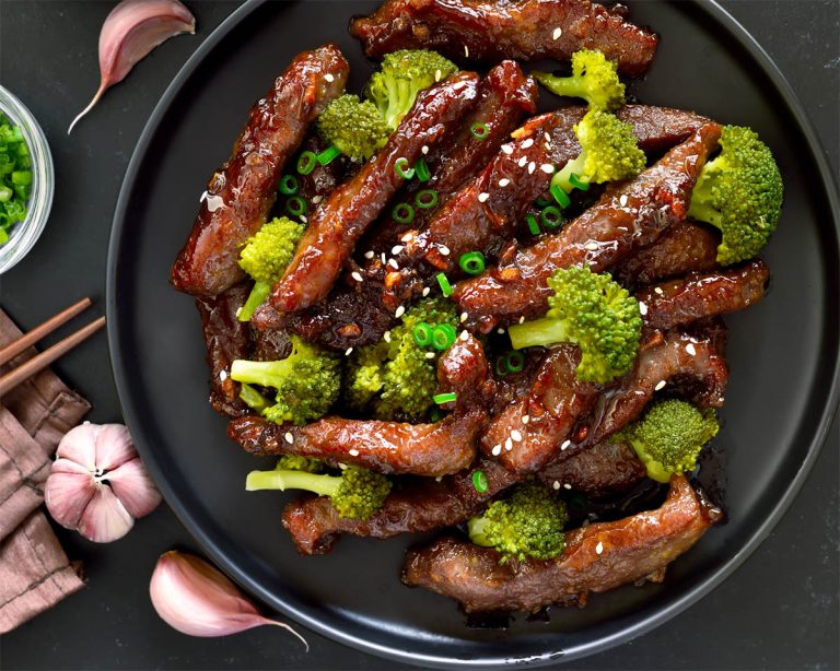 A black plate brimming with glazed beef strips and crisp broccoli florets, garnished with sesame seeds and chopped green onions for a delightful Paleo feast. Nearby, garlic cloves and chopsticks rest on a dark background.