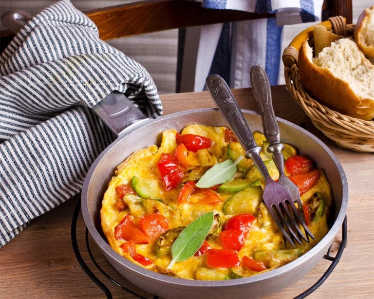 A colorful breakfast frittata with veggies like red peppers and zucchini in a skillet, garnished with a sage leaf. Two forks rest in the skillet, ready to dig into this paleo delight. A basket with bread and a striped kitchen towel are in the background on a wooden table.