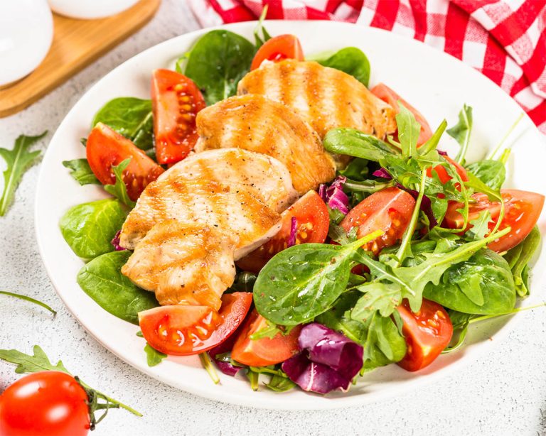 A delicious Paleo-friendly plate of grilled chicken breast sits atop a vibrant salad of mixed greens, including spinach and arugula, garnished with sliced cherry tomatoes. A red checkered napkin and a wooden board with salt and pepper shakers complement the scene in the background.