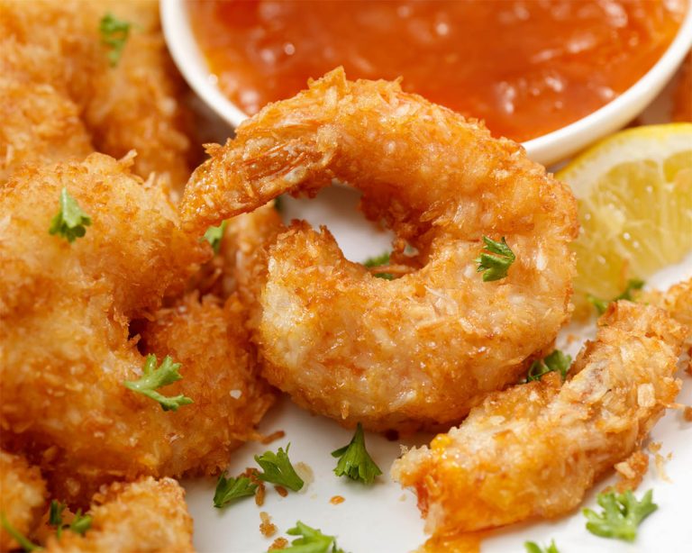 Close-up of crispy coconut-breaded shrimp garnished with fresh parsley, served with a slice of lemon and a bowl of red dipping sauce in the background.