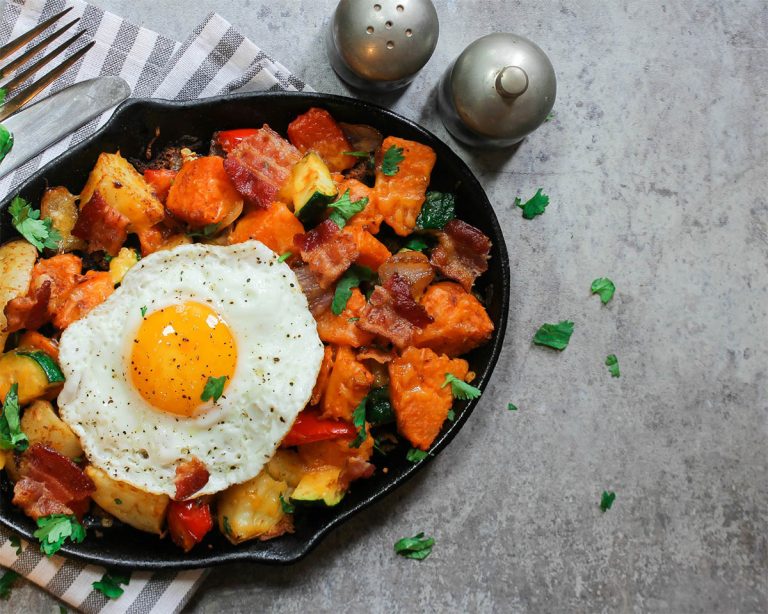 A skillet brimming with a delectable breakfast hash, starring a sunny-side-up egg atop crispy potatoes, sweet potatoes, and bacon. Kale adds a touch of green amidst the herbs. Fresh cilantro garnishes this feast, while a striped napkin and salt and pepper shakers complete the scene.