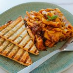 A green plate showcases baked penne pasta in tomato sauce, epitomizing Italian cuisine, garnished with a small basil leaf. On the side, two slices of grilled bread complement the dish. A fork rests invitingly next to the pasta.