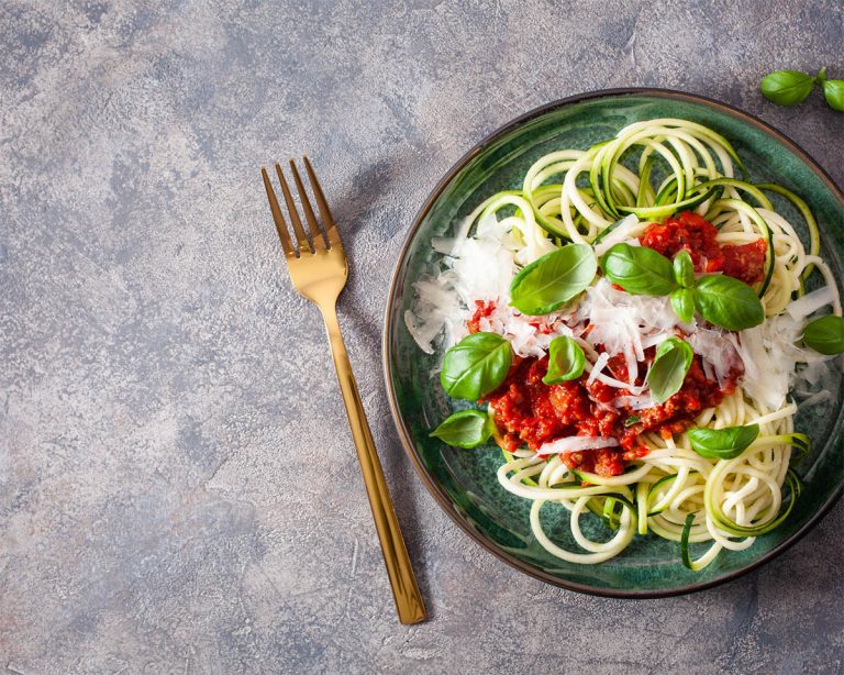 A plate of zucchini noodles topped with rich meat sauce, grated cheese, and fresh basil leaves. A gold fork lies beside the plate on a textured gray surface.