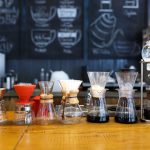 A variety of the best coffee makers for 2024 are displayed on a wooden counter in a café. Behind them, a chalkboard shows coffee-themed drawings and instructions. The setup includes different shapes and sizes of coffee drippers and flasks.