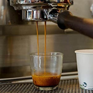 coffee Espresso machine pouring coffee into a clear glass, with a white paper cup nearby. The rich, dark coffee boasts a creamy crema on top. Set against a metallic background that hints at professional barista finesse, it's the perfect start before diving into paleo and vegan recipes.