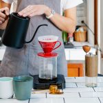 A person wearing a gray apron is using a red pour-over coffee maker to brew coffee. The setup includes a black kettle, a scale, a manual grinder, and two mugs on the tiled counter.