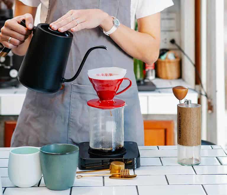 A person wearing a gray apron is using a red pour-over coffee maker to brew coffee. The setup includes a black kettle, a scale, a manual grinder, and two mugs on the tiled counter.