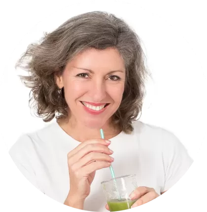 A woman with shoulder-length gray hair smiles while holding a glass of green juice, sipping through a straw. Dressed in a white shirt, she embodies the comfort and simplicity of home, set against a plain white backdrop.