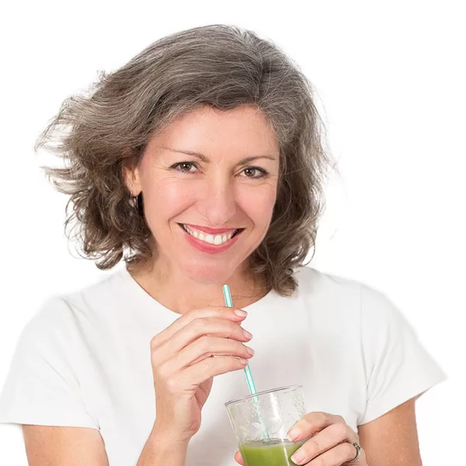 Trish Tucker May 1x1 A woman with shoulder-length gray hair smiles warmly, holding a glass of green juice with a straw, embodying the comfort of home. She is wearing a white top and stands against a serene white background.