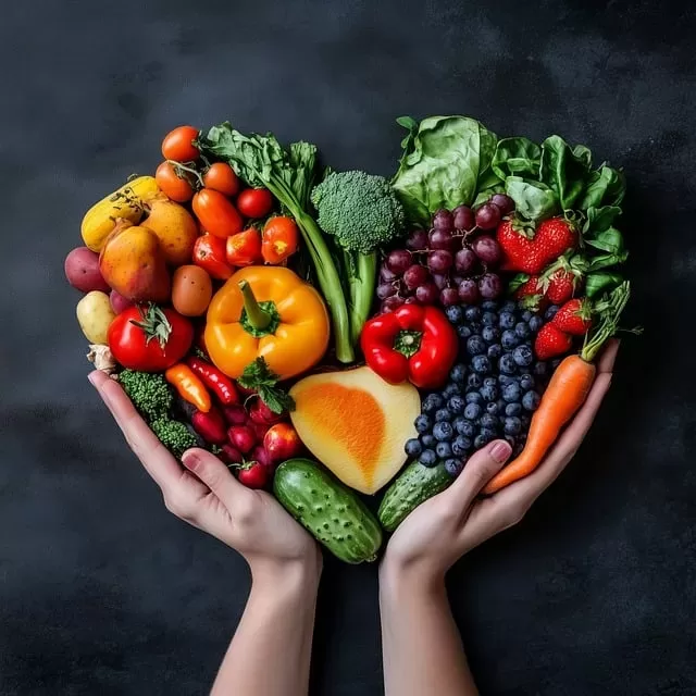 Hands gently cradle a heart-shaped assortment of fresh fruits and vegetables, like bell peppers, carrots, and berries, embodying the vibrant colors of a paleo and vegan lifestyle against a dark background.