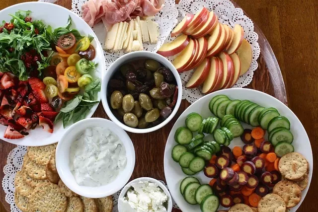 A diverse platter of assorted snacks including sliced apples, various cheeses, cured meats, olives, colorful cherry tomatoes, sliced cucumbers, carrots, leafy greens, crackers, and small bowls of tzatziki and soft cheese showcases the delightful difference in flavors on a wooden table.