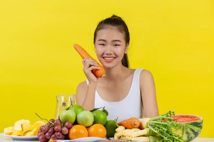 A woman in a white tank top smiles while holding a carrot, embracing her journey towards optimal nutrition. She is seated at a table with assorted fruits and vegetables, including grapes, apples, watermelon, and bananas, against a bright yellow background.