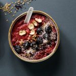 An acai bowl topped with granola, blueberries, banana slices, seeds, and coconut flakes. It rests on a dark surface with small bowls of seeds and berry powder nearby.