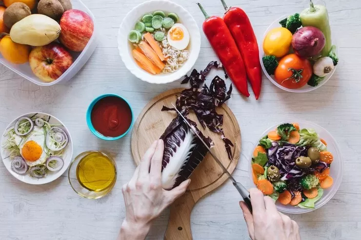 A person embraces the Paleo lifestyle by chopping radicchio on a wooden board surrounded by bowls of fresh vegetables, such as broccoli, carrots, and cucumbers. Nearby are red peppers, fruit, and small bowls containing olive oil and tomato sauce on a light wooden table.