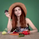 A woman with long red hair wearing a straw hat sits at a table laden with vegetables, including tomatoes, cucumber, carrot, garlic, and avocado. Holding an avocado and gazing thoughtfully to the side, she ponders the difference between paleo and vegan lifestyles.