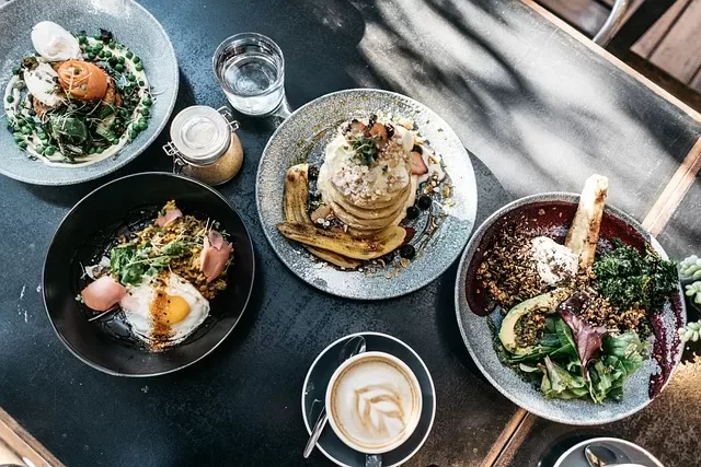 A dark table is set with plates of food, including a salad with greens and grains, a poached egg dish, and a cappuccino in a white cup. Pancakes are topped with bananas in true paleo diet style. A small glass of water and a jar with a lid complete the scene.