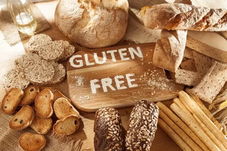 A variety of health-focused, gluten-free breads and crackers are displayed on a wooden surface. In the center, "GLUTEN FREE" is written on a wooden board surrounded by delicious loaves, rolls, and breadsticks.