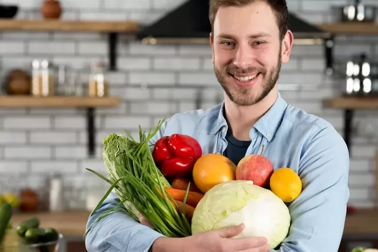 A smiling man stands in a kitchen holding a variety of fresh produce, perfect for paleo and vegan diets, including cabbage, red pepper, oranges, and green onions. The background features shelves with jars and a modern tiled wall, reminiscent of kitchens found across the USA and UK.