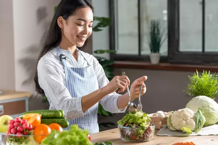 A woman in a striped apron prepares a fresh salad in her bright kitchen, embracing a paleo and vegan diet. She smiles while using utensils to mix greens, surrounded by various vegetables on the table, including radishes, cucumbers, and cauliflower.