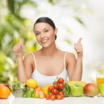 A woman smiling and showing thumbs up sits behind a table filled with fresh fruits and vegetables, including apples, tomatoes, mangoes, lettuce, carrots, berries, and a glass of orange juice. A bright, leafy background highlights her commitment to healthy habits and optimal nutrition.