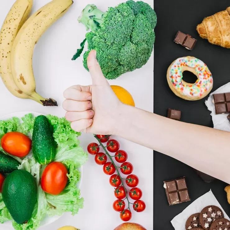 A variety of foods, including bananas, broccoli, and tomatoes for optimal nutrition alongside indulgent treats like croissants and chocolate, are arranged on a half white, half black background. A hand with a thumbs up gesture in the center symbolizes balance in weight management.