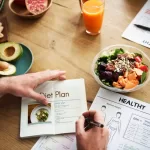 A person writing in a diet plan journal at a wooden table, surrounded by nourishing foods like avocado, nuts, a salad bowl, and a glass of orange juice. Nearby are BMI charts and nutritional info papers reflecting their commitment to a paleo vegan lifestyle.
