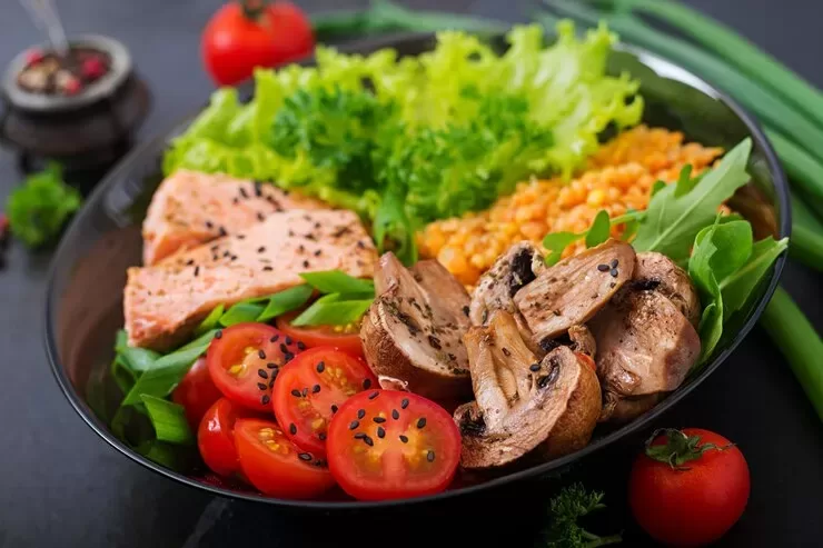 A vibrant paleo dish featuring grilled salmon, cherry tomatoes, sautéed mushrooms, lettuce, green onions, and a serving of lentils, garnished with sesame seeds and beautifully displayed on a black plate.