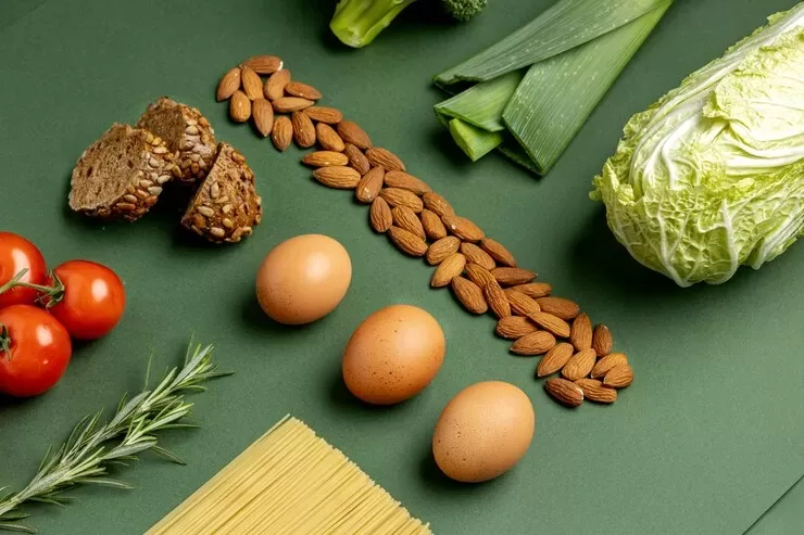 A flat lay of fresh ingredients, highlighting a wealth of options for both paleo and keto enthusiasts, features tomatoes, rosemary, whole grain bread, almonds, eggs, spaghetti, leeks, broccoli, and a leafy cabbage on a vibrant green surface.