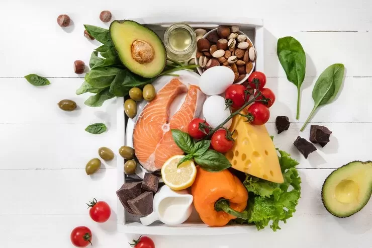 A variety of foods, including salmon, cheese, avocado, and eggs—all essentials for optimal nutrition—alongside nuts, leafy greens, cherry tomatoes, olives, a bell pepper, lemon slices, and dark chocolate are artfully arranged on a white surface.