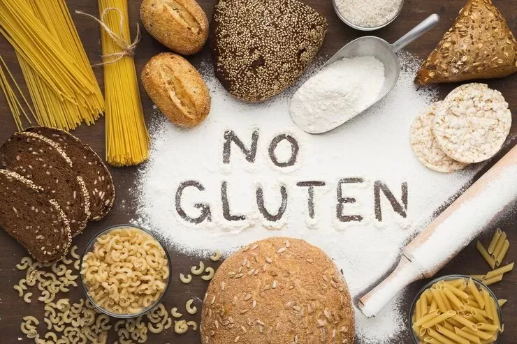 An assortment of health-conscious, gluten-free foods, including bread, pasta, and rolls, is arranged around flour with "NO GLUTEN" written on it. A rolling pin and scoop of flour are nearby, set on a wooden surface.