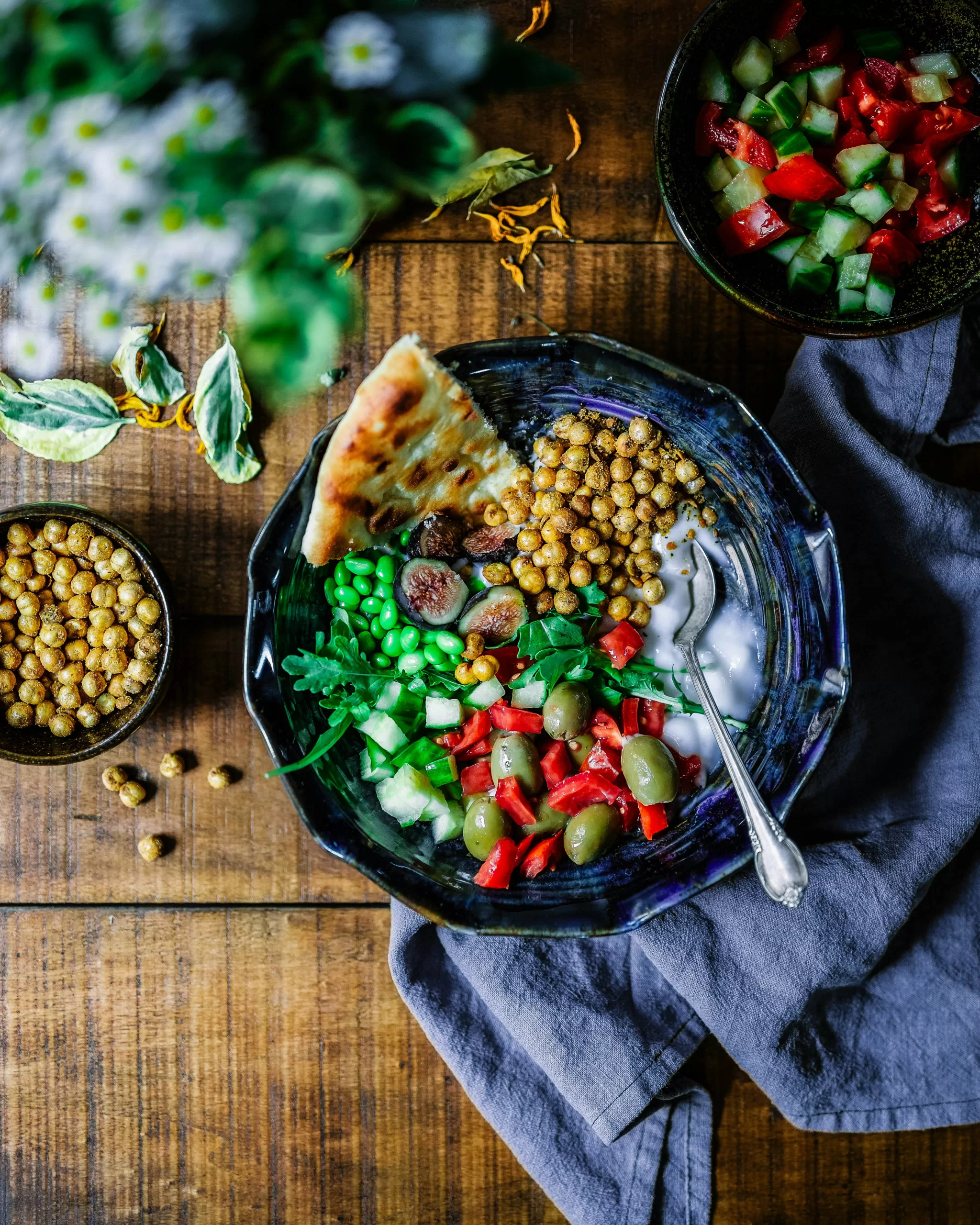 A colorful salad bowl on a wooden table, embraced by vegan influencers, features chickpeas, olives, peas, diced tomatoes, cucumber, greens, and a wedge of flatbread. Nearby lie extra bowls of chickpeas and salad with a gray cloth napkin beside the main plate.