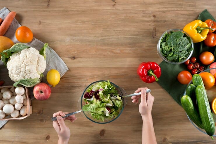 Hands mix a vibrant salad in a glass bowl on a wooden table, embodying the essence of Vegan Life. Surrounding the bowl are fresh vegetables and fruits like peppers, oranges, cauliflower, apples, and mushrooms, creating a colorful and healthy spread fit for any 2024 guide to clean eating.