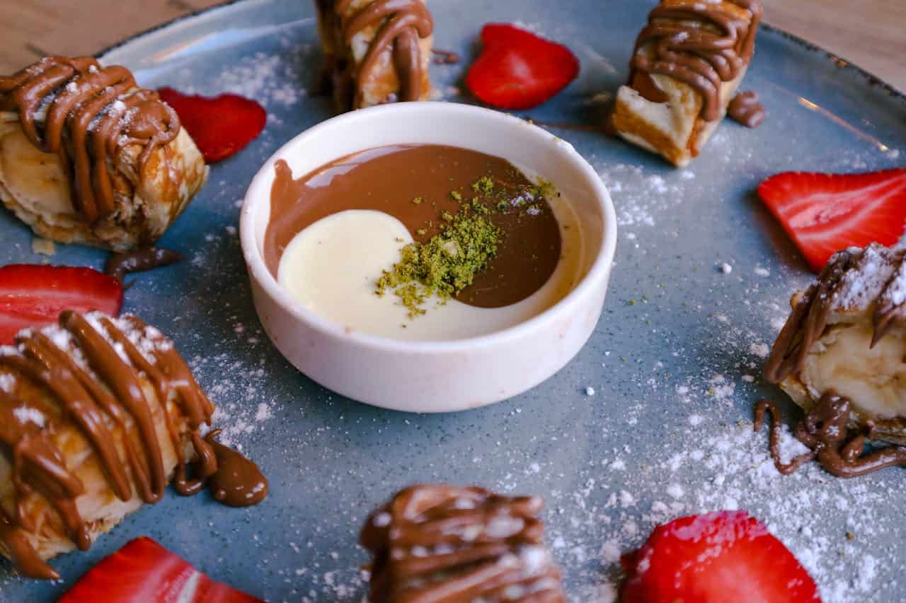 A beautifully arranged dessert plate featuring sliced strawberries, pastry-wrapped bananas drizzled with chocolate, and a bowl of swirled milk and dark chocolate sauce garnished with a sprinkle of green pistachio dust in the center. The plate is dusted with powdered sugar, adding an elegant touch.