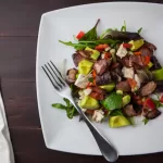 A square white plate holds a salad with mixed greens, avocado, red bell peppers, diced chicken, and pieces of crispy bacon reminiscent of French cuisine. A fork rests on the plate. The background features a dark wooden table and a light-colored cloth.
