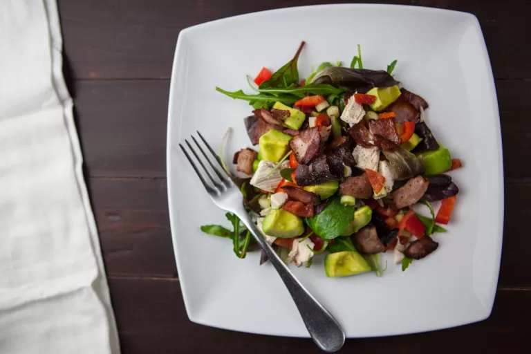 A square white plate holds a salad with mixed greens, avocado, red bell peppers, diced chicken, and pieces of crispy bacon reminiscent of French cuisine. A fork rests on the plate. The background features a dark wooden table and a light-colored cloth.