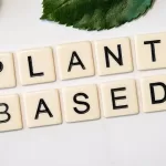 Scrabble tiles spell "PLANT BASED" on a white surface surrounded by green leaves, capturing the essence of vegan living—a key thing to know for those exploring how to become vegan.