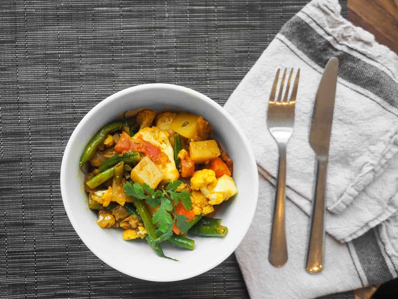 A white bowl filled with vibrant vegan vegetable curry, featuring green beans, cauliflower, potatoes, and carrots, garnished with cilantro. The bowl is placed on a black woven placemat next to a folded napkin with a fork and knife—a delightful dish if you decide to become vegan.