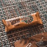 A vegan banana and hemp raw bar in a golden wrapper, placed on a textured, woven surface with shades of teal, brown, and white. The packaging highlights features like "preservative-free," "cholesterol-free," and "no artificial flavors or colors." A decorative earring rests on a wooden tray nearby, adding an artistic touch to the composition.