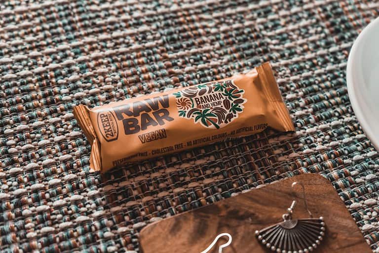 A vegan banana and hemp raw bar in a golden wrapper, placed on a textured, woven surface with shades of teal, brown, and white. The packaging highlights features like "preservative-free," "cholesterol-free," and "no artificial flavors or colors." A decorative earring rests on a wooden tray nearby, adding an artistic touch to the composition.