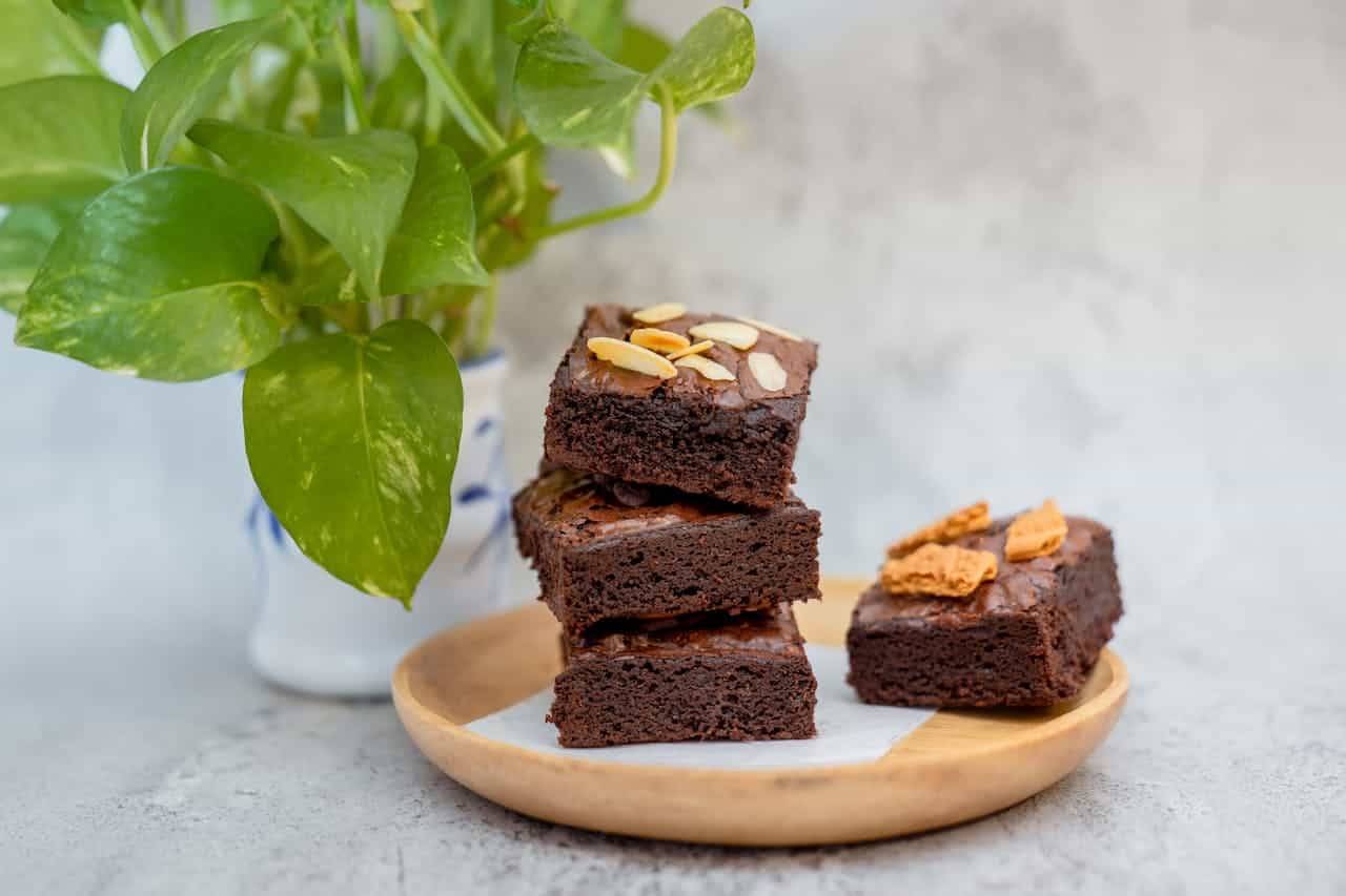 A wooden plate with a stack of rich, fudgy chocolate brownies, topped with sliced almonds and biscuit pieces for added texture. The brownies are placed on a parchment sheet, and a vibrant green potted plant is in the background, adding a fresh, natural touch to the scene. 