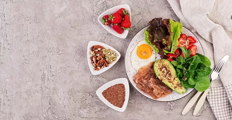 A breakfast plate perfect for paleo vs keto enthusiasts features crispy bacon, a sunny-side-up egg, sliced avocado, lettuce, and cherry tomatoes. Nearby bowls offer strawberries, mixed nuts, and flaxseeds. A folded napkin and fork rest on the textured gray surface to complete the setting.