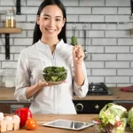 In a kitchen filled with vibrant tomatoes, peppers, and cabbage, a woman holds up a bowl of leafy greens. As she stands before her tablet on the wooden counter, she contemplates the age-old paleo vs keto debate amidst the fresh array surrounding her.