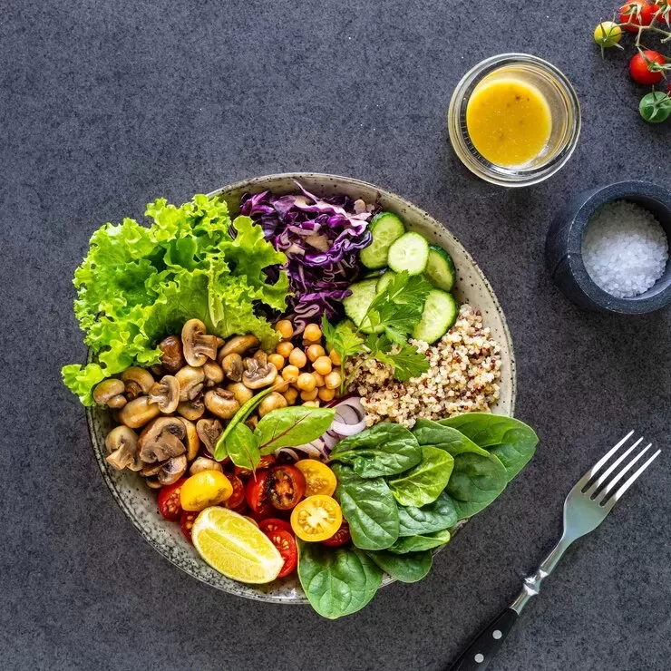 A vibrant paleo vegan salad bowl with quinoa, leafy greens, cherry tomatoes, chickpeas, mushrooms, and cucumber slices garnished with lemon wedges. A small bowl of dressing and a dish of salt are nearby on a dark surface.