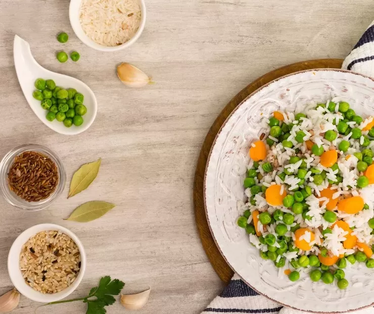 A plate of rice mixed with peas and carrots rests on a wooden surface, showcasing easy vegan flavors. On the side are small dishes with white rice, lentils, peas, and garlic cloves, perfect vegan substitutes. A striped cloth lies partially under the plate.