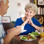 An elderly woman in a blue shirt smiles while sitting at a table with salad and orange juice, enjoying a lively chat about the benefits of a vegan diet with an elderly man in a plaid shirt holding his coffee cup. Shelves filled with books and photos provide the perfect backdrop to their discussion.