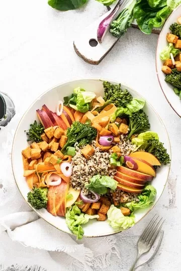 A colorful salad bowl featuring quinoa, diced tofu, sliced peaches, broccoli, lettuce, carrots, and red onion rings is arranged neatly on a white plate. A wooden cutting board and fresh greens are in the background—highlighting easy vegan substitutes for common foods.