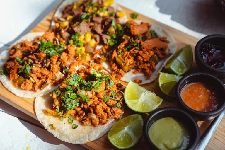 A wooden platter showcases a delightful Mexican cuisine featuring four tacos loaded with seasoned meat, cilantro, and diced vegetables. Slices of lime accompany the feast, along with three small bowls of vibrant sauces, embodying the rich flavors of Mexican food.