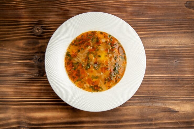 A white bowl filled with pizza soup featuring diced vegetables and herbs, perfect for those on a Paleo diet, sits on a wooden table background.