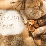 A variety of gluten-free breads and grains are displayed on a wooden surface. Flour on the table spells out "gluten free." The health-conscious assortment includes crusty bread, crackers, and uncooked pasta.