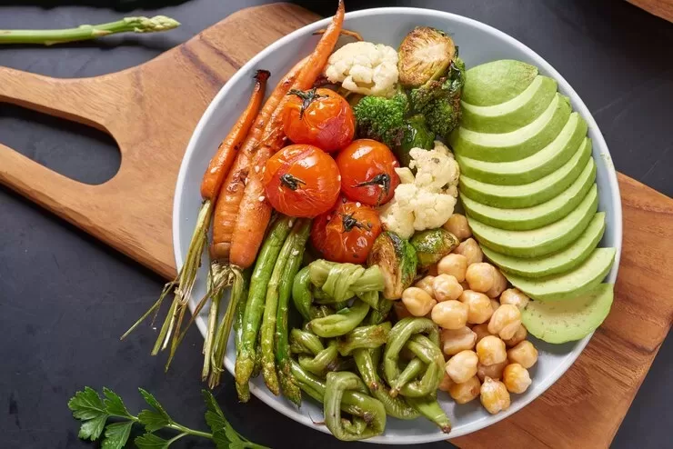 A colorful plate of common foods like roasted cherry tomatoes, carrots, green beans, bell peppers, Brussels sprouts, broccoli, cauliflower, and chickpeas. The dish is easy to prepare and features sliced avocado as a delightful vegan substitute on a wooden board.